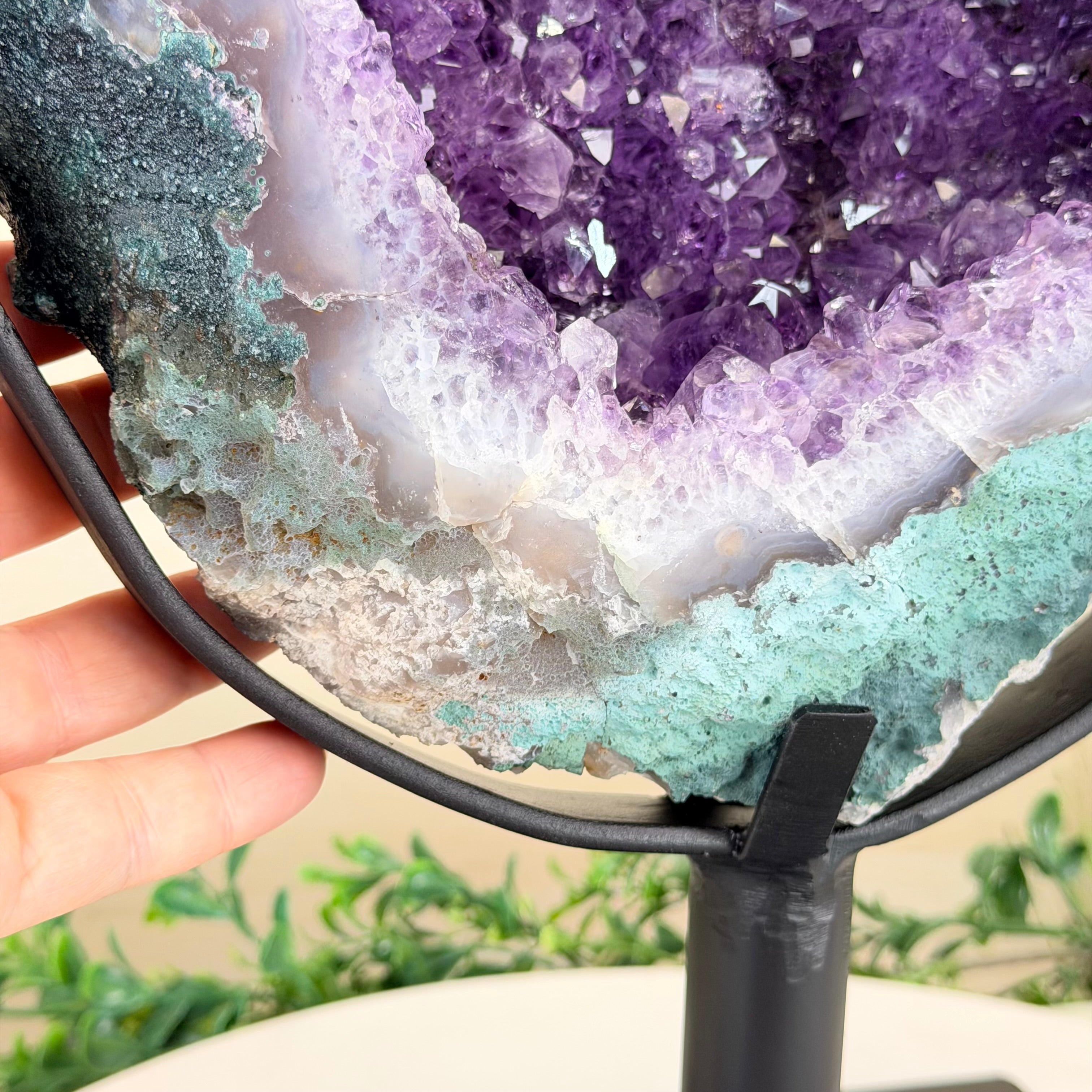 Close-up of a multicolored crystal formation held by a hand with a blurred green plant background.