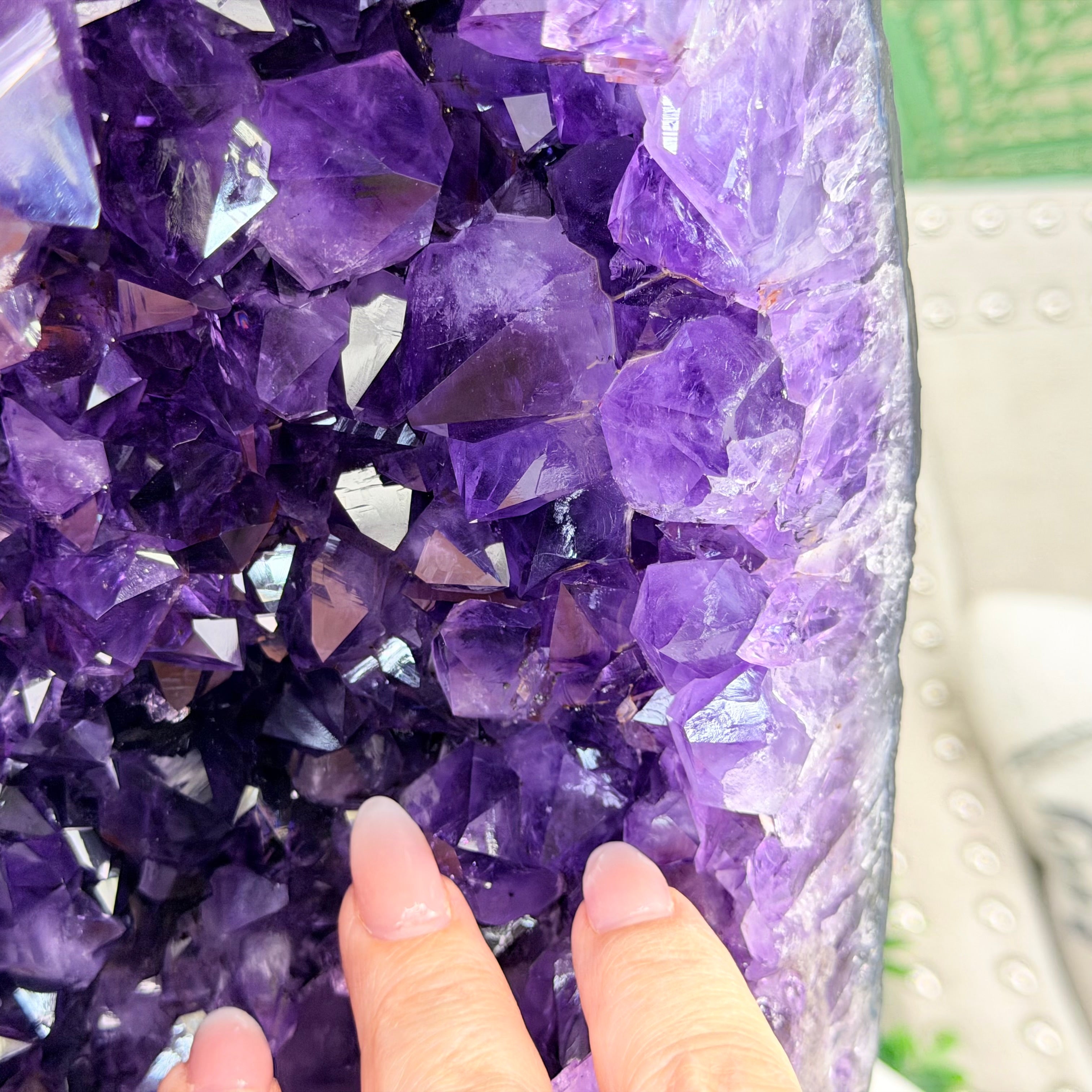 A close-up of a purple amethyst crystal geode cluster with a person's fingers pointing at it, set against a blurred background.
