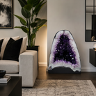 A large freestanding amethyst geode wall decor piece with deep purple crystal clusters and white matrix edges, displayed in a modern living room next to a beige sofa and a potted plant.
