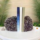 Two large crystal geode bookends made of dark purple amethyst are displayed on a white round table, holding three upright books. A small card reading BRAZILGEMS is visible on the right side.