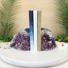 Two crystal geode bookends made from purple amethyst and clear quartz, holding up three books on a white round tray. The background features blurred green palm leaves and a beige wall.