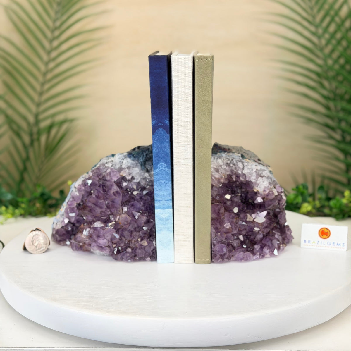 Two crystal geode bookends made from purple amethyst and clear quartz, holding up three books on a white round tray. The background features blurred green palm leaves and a beige wall.