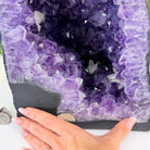 A large purple amethyst geode specimen with a black matrix base, shown with a person's hand resting along the bottom edge for scale.