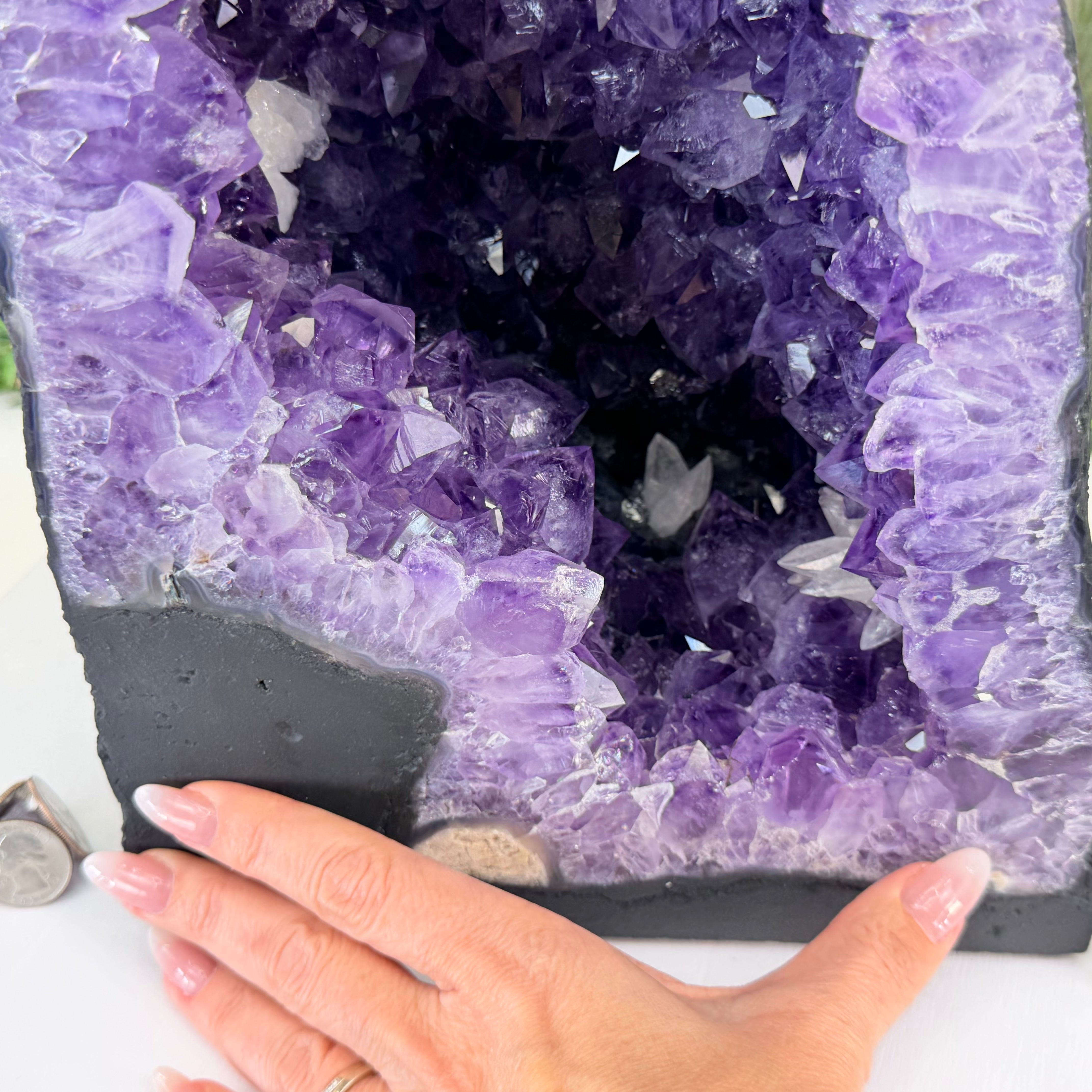 A large purple amethyst geode specimen with a black matrix base, shown with a person's hand resting along the bottom edge for scale.