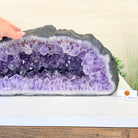 Large amethyst geode with a hand on a white surface, branded 'BrazilGems'.