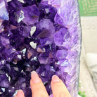 A close-up of a purple amethyst crystal geode cluster with a person's fingers pointing at it, set against a blurred background.