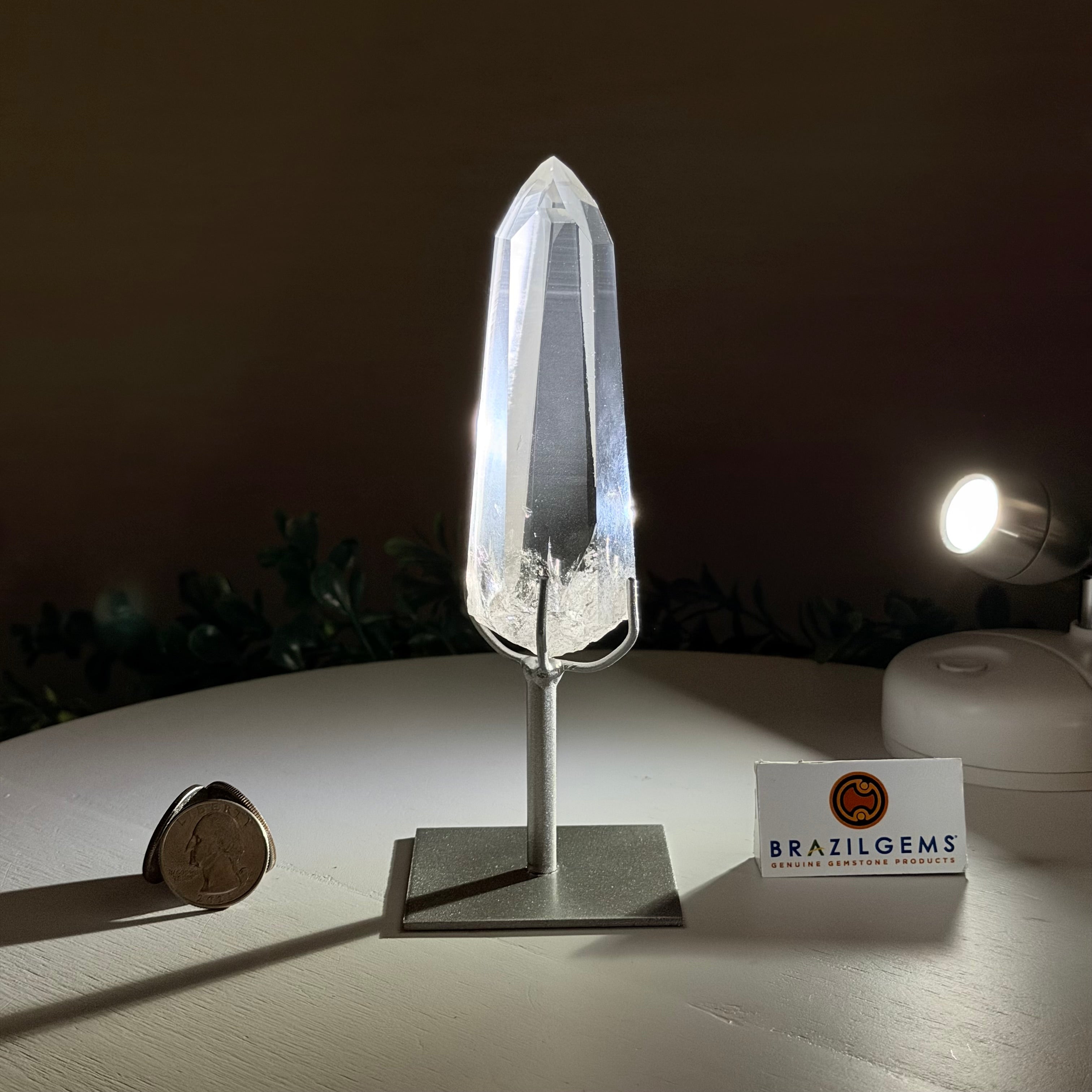 Crystal on a stand with a coin for scale, next to a 'BrazilGems' card on a dark background.